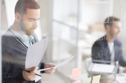 Side view portrait of handsome bearded man reading documents standing behind glass wall while working in office